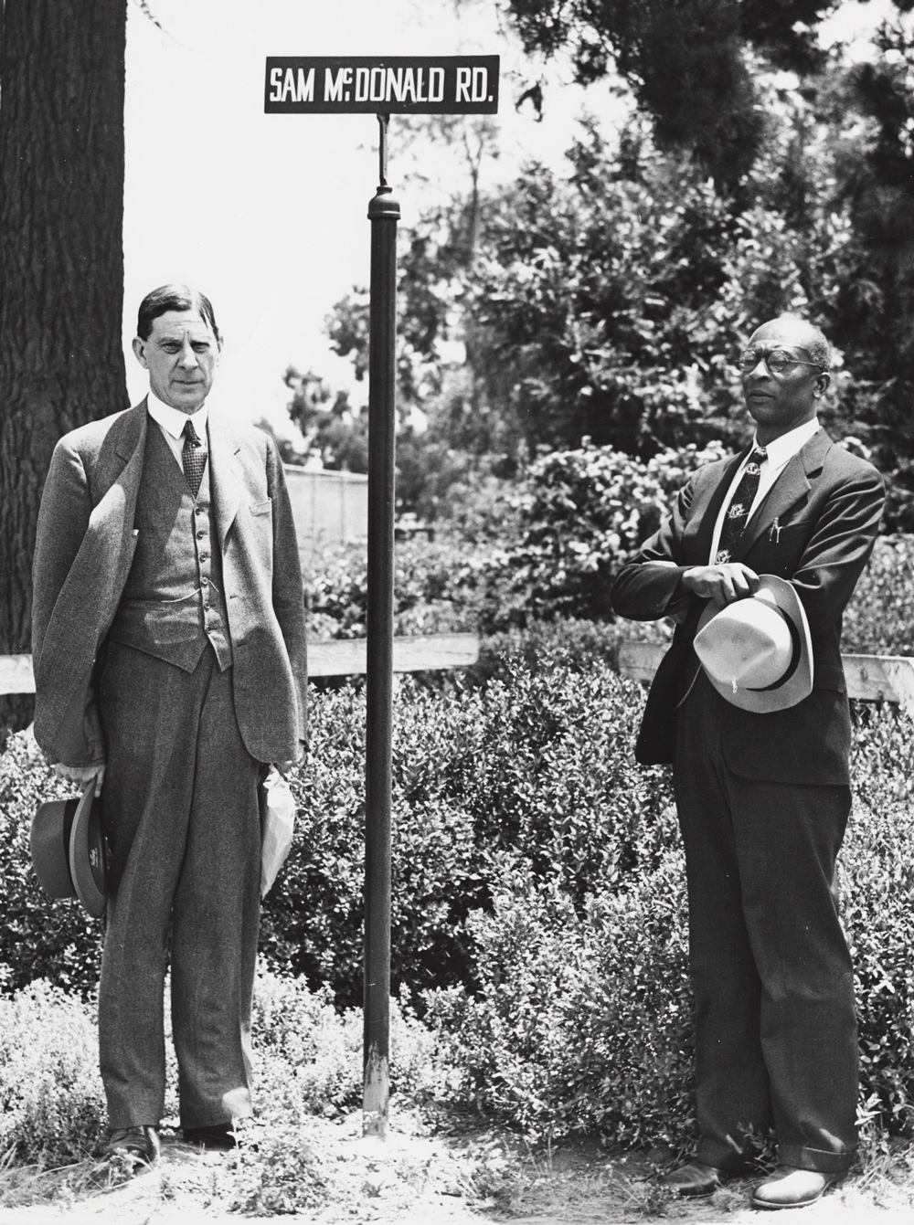 Black and white photo of Ray Lyman Wilbur and Sam McDonald, in suits and ties, standing by the street sign for Sam McDonald Way.