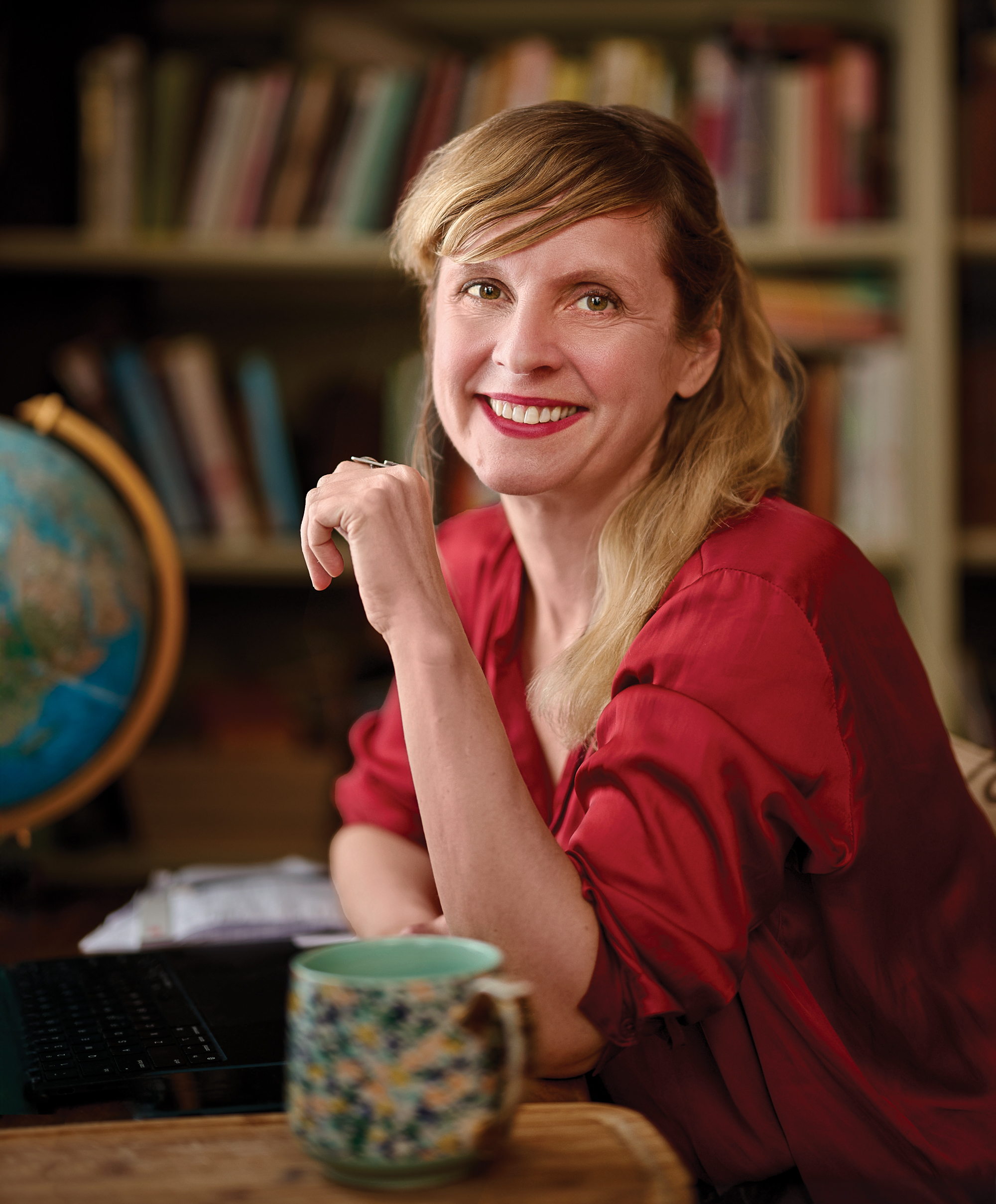 Leigh Newman at a desk with a coffee cup on a table and a globe and bookshelf in the background