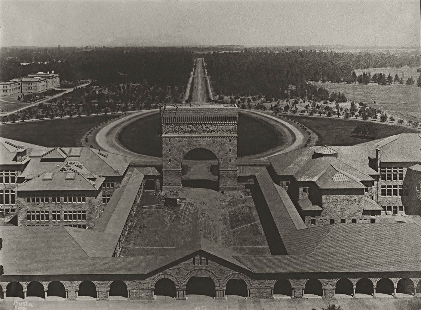 An early picture of the entrance to Stanford's Main Quad. Past the Quad's border wall is a sizable courtyard with buildings on both sides. At its entrance is a large stone arc. Beyond that is a large oval road which converges some ways down into a single road leading far off into a forest.