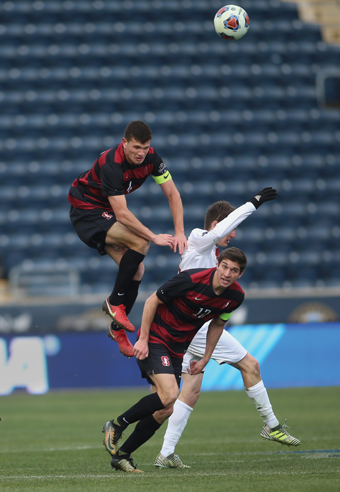 Three male soccer players on the field, two on the Stanford team. They're all closely packed together with one Stanford player jumping over the other two.