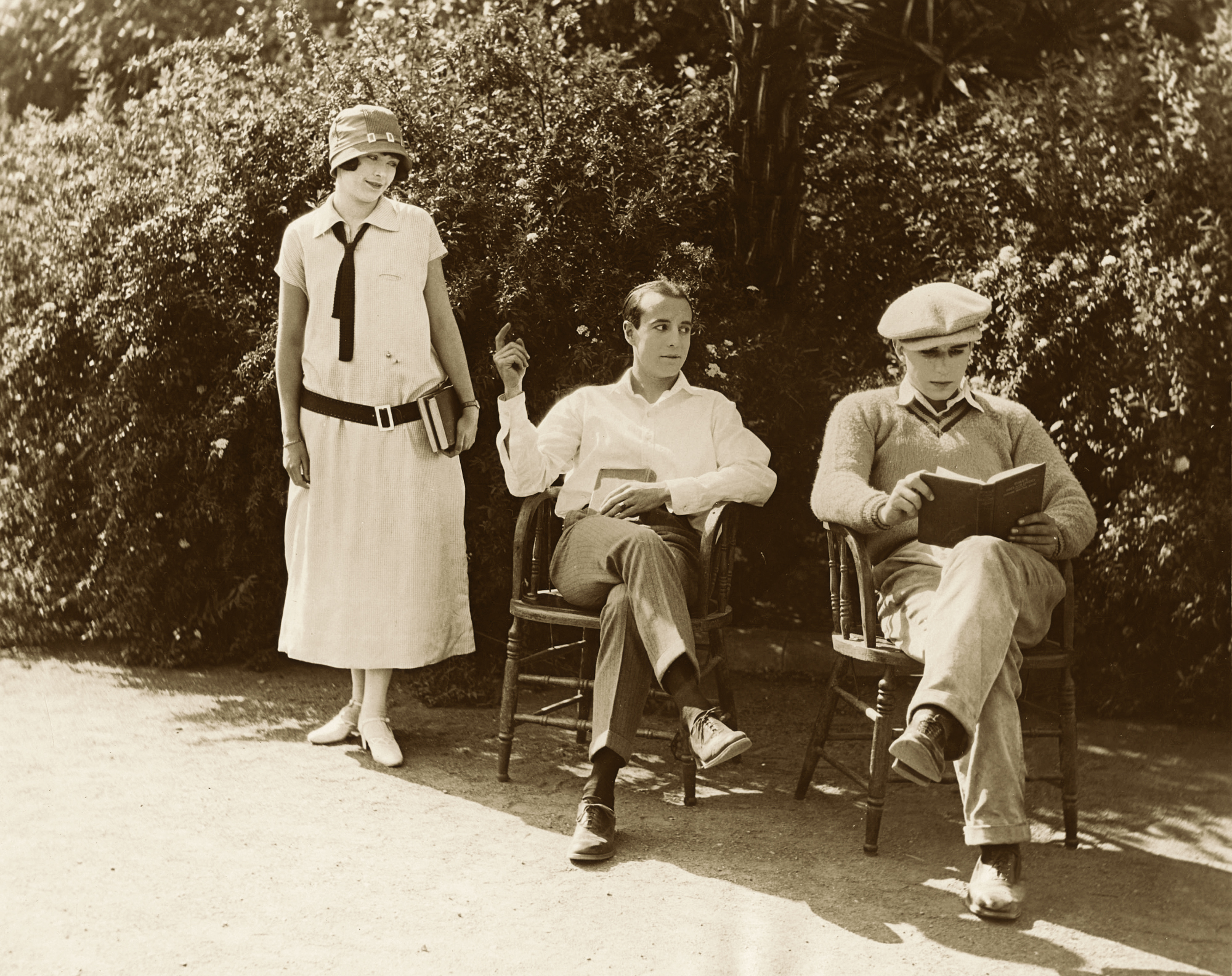 A woman standing next to two men sitting in chairs, each with books.