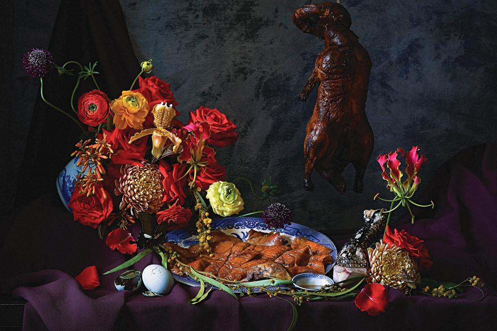 Flowers on a table along with a plate of fish and a hanging roasted duckling